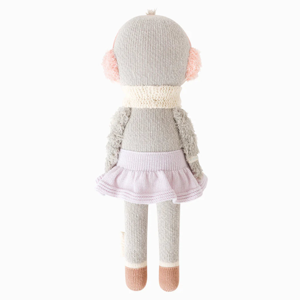 A fair trade toy, Cuddle + Kind Skylar The Penguin is a handmade knitted stuffed animal resembling a penguin, shown from the back, wearing a light purple skirt, cream-colored scarf, and pink earmuffs, with brown feet and gray fuzzy arms.