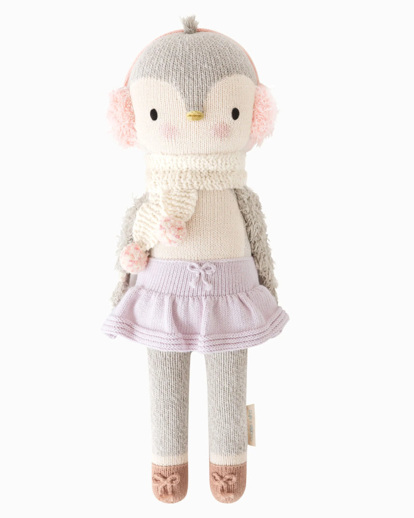 Cuddle + Kind Skylar The Penguin is a handmade knit cotton doll with gray and white coloring, pink earmuffs, a white scarf with pink accents, lavender skirt, taupe shoes, and pink cheeks—a charming fair trade plush toy.