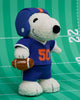 The Peanuts Snoopy - American Football plush from Bon Ton Toys features Snoopy in a blue football uniform with number 50, holding a brown football and standing on a green field with white yard lines.