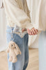 Two people stand close, holding hands. One wears a cream sweater and blue jeans, accessorized with a silver purse and the Snuggle Bunnies - Penelope Keyring in Caramel. The softly lit, neutral background creates a cozy vibe.