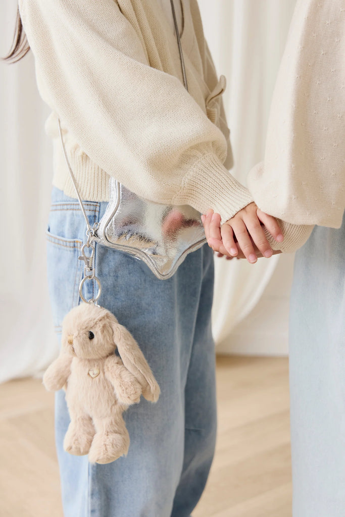 Two people stand close, holding hands. One wears a cream sweater and blue jeans, accessorized with a silver purse and the Snuggle Bunnies - Penelope Keyring in Caramel. The softly lit, neutral background creates a cozy vibe.