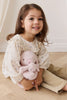 A young girl with long brown hair, dressed in a cream floral blouse and beige pants, sits on a wooden floor, smiling as she hugs her Snuggle Bunnies—Frankie the Hugging Bunny and Blossom—soft plush toys that bring comfort and joy.
