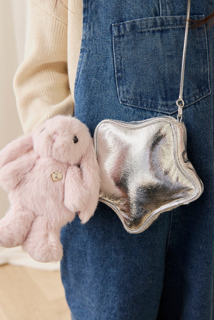 A person in denim overalls holds a plush pink bunny and wears a shiny silver star-shaped purse adorned with the Snuggle Bunnies - Penelope Keyring - Blossom on the shoulder strap.