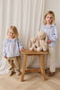 Two blonde-haired children in light blue tops and beige pants stand by a wooden stool holding Snuggle Bunnies - Penelope - Caramel plush toys. A white curtain in the background completes this charming scene.