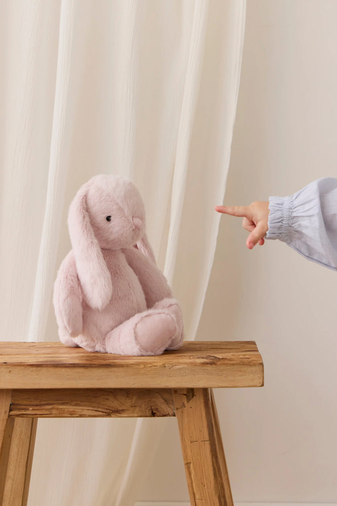 The Snuggle Bunnies - Penelope - Blossom, a soft pink plush bunny, sits on a wooden bench before a sheer white curtain as a child's blue-sleeved hand points at it from the right.