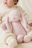 A baby dressed in a fuzzy cream outfit sits on a light surface, holding the Snuggle Bunnies - Penelope - Blossom, a soft light pink plush bunny with long ears. The baby's face is partially visible.