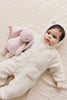 A baby in a cozy white hooded onesie with bear ears lies on a cream blanket, hugging the Snuggle Bunnies - Penelope - Blossom plush stuffed animal in light pink.