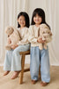 Two young girls in cream sweaters and light blue jeans smile while sitting together on a wooden floor, each holding Snuggle Bunnies - Frankie the Hugging Bunny - Caramel plush toys, posing in front of a white curtain.