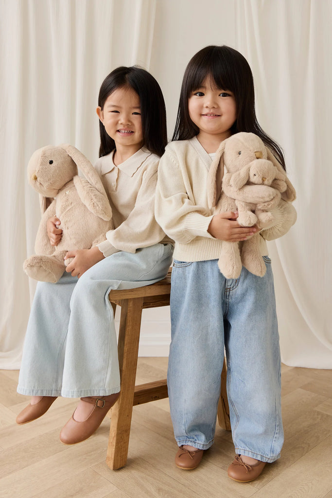 Two young girls in cream sweaters and light blue jeans smile while sitting together on a wooden floor, each holding Snuggle Bunnies - Frankie the Hugging Bunny - Caramel plush toys, posing in front of a white curtain.