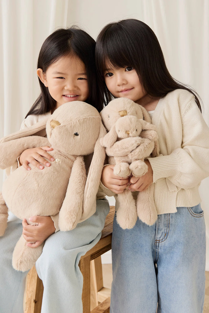 Two young girls with long dark hair in light sweaters and blue jeans sit together on a wooden bench, smiling as they hold Snuggle Bunnies – Frankie the Hugging Bunny in caramel. The background is light.