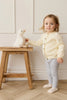 A young child with light hair, in a cream sweater and gray leggings, stands indoors touching Snuggle Bunnies - Rose The Duck, a super soft plush toy on a wooden stool against a light-colored wall.