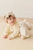A baby in a cream knit sweater with a big matching headband lies on their stomach next to a super soft Snuggle Bunnies - Rose The Duck plush toy on a light-colored floor.