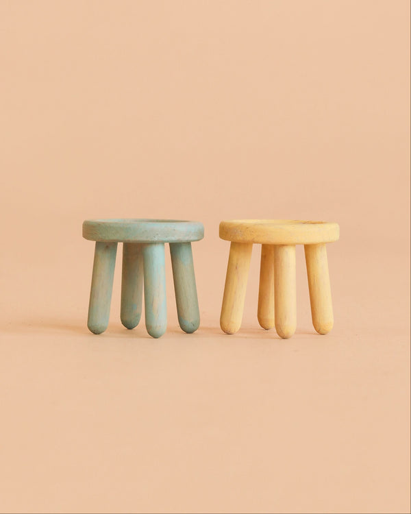 Two small Maileg Stool Set, Mouse stools—one in mint and one in light yellow—crafted from beech wood with a vintage paint finish, sit side by side on a peach-colored surface against a matching peach background.