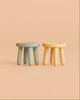 Two small, round stools with chunky legs—one blue and one yellow—sit side by side against a plain peach background, perfect as part of the Maileg Cozy Christmas Corner collection for your mouse furniture or dollhouse accessories.