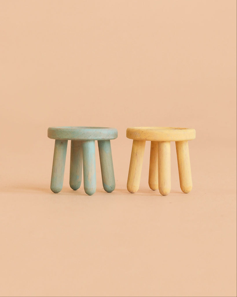 Two small, round stools with chunky legs—one blue and one yellow—sit side by side against a plain peach background, perfect as part of the Maileg Cozy Christmas Corner collection for your mouse furniture or dollhouse accessories.