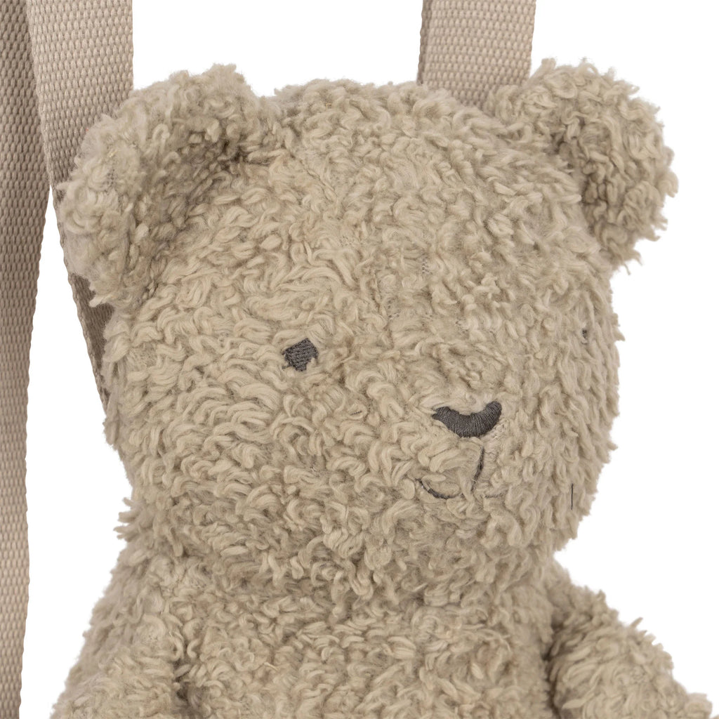 A fluffy, light brown teddy bear with curly organic sherpa fur and simple stitched facial features is positioned in front of a white background. A beige fabric strap is visible behind the bear, hinting at its function as an adorable Konges Slojd | Teddy Sherpa Bear Backpack. The teddy has round ears and a slightly smiling expression.