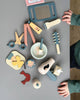 A child's hands reaching towards a collection of colorful wooden toys and craft supplies arranged neatly in a Hair Salon. Items include a drawing pad, scissors, a paintbrush, and decorative shapes.