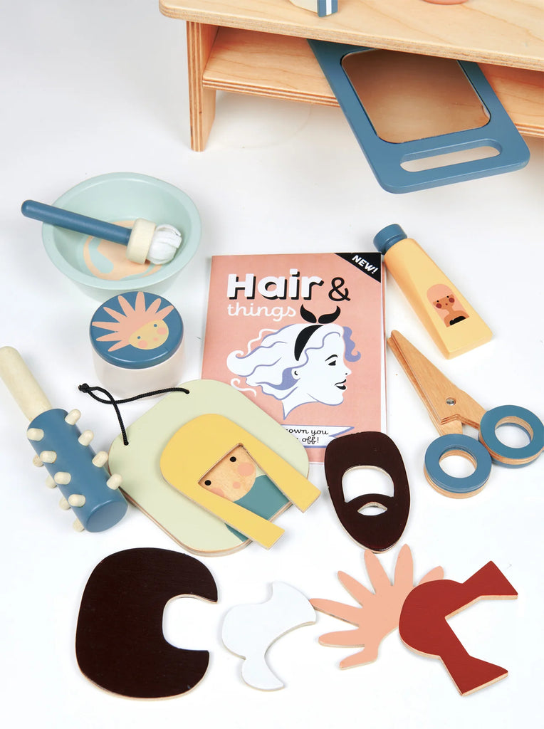 Assorted wooden baby toys, including a puzzle, teether, and the Hair Salon book, arranged on a white surface against a wooden backdrop.