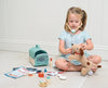 A young girl in a blue dress pretends to give a checkup to a stuffed dog toy, using the Animal Rescue Vet Case’s toy stethoscope and colorful veterinary play set. Various pretend medical supplies from the Animal Rescue Vet Case are spread on the floor beside her, along with a pet carrier toy, creating an imaginative and playful vet scene.
