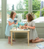 Two young girls sit at a small table indoors, using their Animal Rescue Vet Case to pretend to care for a toy dog. Toy cups and utensils are spread out on the table, and a pet carrier toy sits nearby. Sunlight pours in through large windows, revealing a green garden outside.