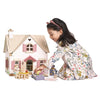 A young girl plays with the Cottontail Cottage Dollhouse featuring a natural wood roof, arranging miniature figures and retro style furniture. She wears a floral dress and a pink headband, focused and engaged in her play.