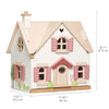 A Cottontail Cottage Dollhouse featuring a beige exterior with pink shutters, trim, and a front door. The house displays a clock and a small white rabbit by the front wall. It includes measurement annotations on.
