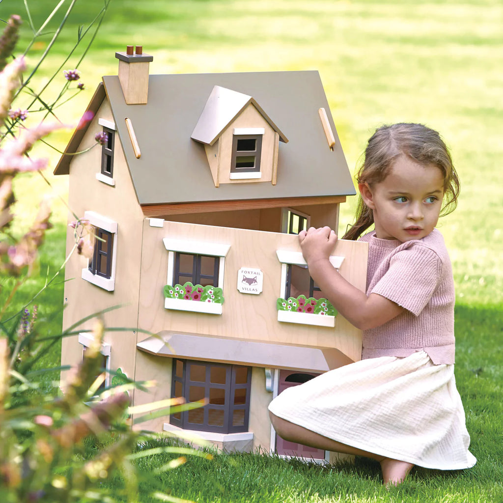 A young girl kneels next to a large, detailed Foxtail Villa Dollhouse in a grassy yard, looking thoughtfully at the camera. The dollhouse has realistic features including windows and flower boxes.