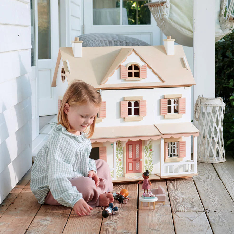 A young girl with blonde hair is sitting on a wooden porch, playing with small dolls in front of a large Humming Bird Dollhouse. The colonial-style home is white with pink window shutters and a detailed front door. The girl is smiling and appears engaged in her playtime, showcasing her miniature furniture collection.