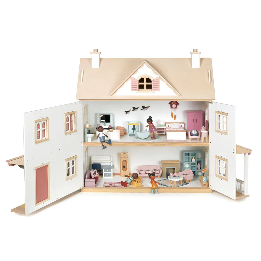 The Humming Bird Dollhouse is a wooden dollhouse with a detailed interior, featuring multiple rooms across two floors. This charming colonial-style home comes furnished with a miniature furniture collection and accessories, and several small, diverse dolls occupy various spaces, engaging in different activities. The dollhouse is open in the front.