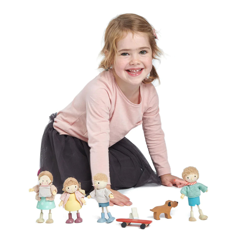 A young girl with a joyful smile, crouching on the floor, playing with four small toy figures including Mr. Goodwood and his Dog, on a white background.
