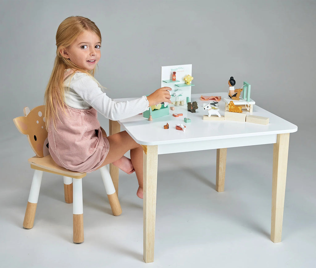 A young girl with blonde hair sits on a deer-themed chair, playing with toy animal figures and her Waggy Tails Dog Salon on a white table with light wooden legs. The background is plain and grey, and she is smiling and engaged in her activity.