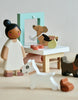 A scene with wooden toy figures, including a person with a bun hairstyle, wearing a white dress, two dogs—one white in the foreground and a brown one jumping through a green frame on the table. The imaginative playtime setup also features two brown dog bowls, inviting fun and stories of various dog breeds in the Waggy Tails Dog Salon.