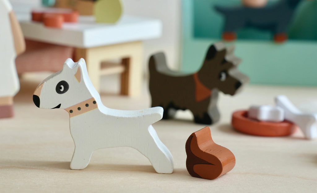 A close-up shot of a small white wooden dog figurine with a collar standing on a light-colored surface. In the background, a small dark brown wooden dog figurine is partially visible. A wooden food bowl and a chew toy are also visible nearby, perfect for imaginative playtime with various dog breeds from Waggy Tails Dog Salon.