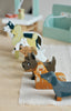A collection of small, colorful wooden dog figures arranged in a line on a light-colored table. The dogs, representing various dog breeds, are painted in shades of brown, black, white, and gray, each with unique markings and features. A Waggy Tails Dog Salon is blurred in the background.