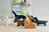A collection of wooden dog figurines, showcasing various colors and dog breeds, are arranged on a table. In the background, there's a shelf with towels, a duck figurine, and grooming supplies. The scene evokes imaginative playtime at Waggy Tails Dog Salon.