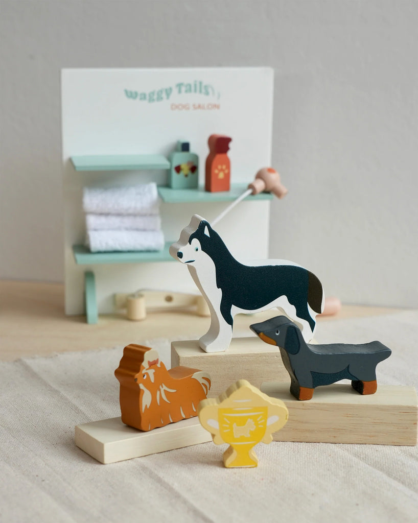A small set of wooden dog figurines, including a Husky, a Dachshund, and a couple of other breeds, are arranged in front of a miniature spa backdrop labeled "Waggy Tails Dog Salon," perfect for imaginative playtime focused on dog grooming. Tiny shelves hold towels and bottles beside a trophy figure.