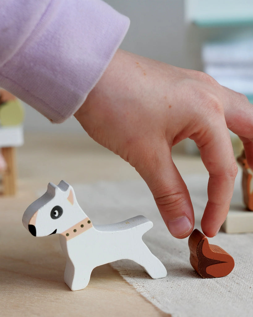 A child's hand reaches for a small, brown wooden figure beside a white wooden dog toy with black details from the Waggy Tails Dog Salon, placed on a beige surface. The child is wearing a light purple sleeve.