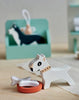 A small wooden toy dog with a white body and tan collar stands next to a red bowl and a white bone. Behind it, there is a green rectangular block featuring another dog illustration with "Waggy Tails Dog Salon" branding and a person sitting. The scene, part of the dog grooming play set, is set on a light-colored surface.