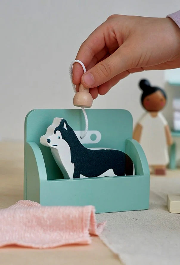 A child's hand is playing with a small toy dog and a bed. The black and white dog from the Waggy Tails Dog Salon rests on the light teal bed. A smaller peg doll figure with a bun hairdo is seen in the background. There's a peach-colored piece of fabric in the foreground.