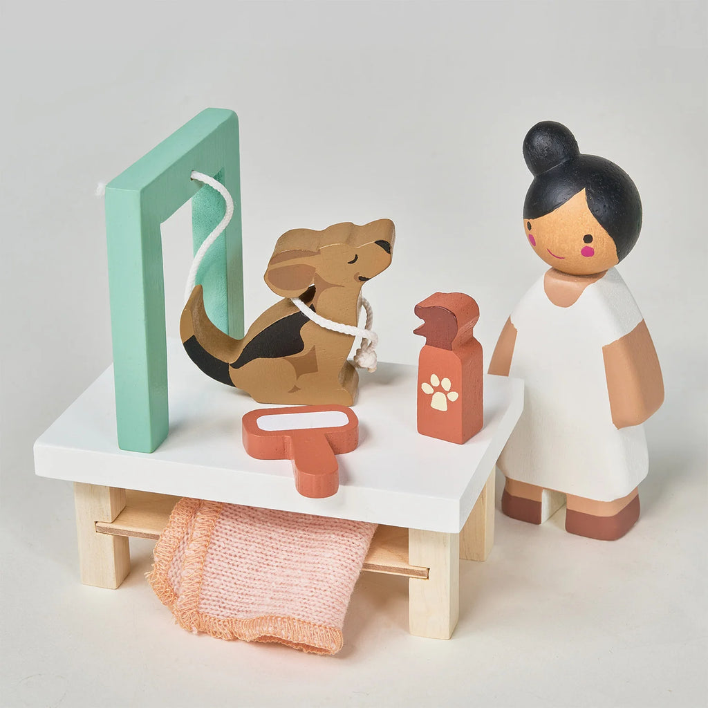 The Waggy Tails Dog Salon encourages imaginative playtime. A brown dog figure, resembling popular dog breeds, is held by a white rope attached to a light green frame. Nearby, a person figurine with a topknot stands next to a bottle with a paw print, pink towel, and comb on a white table.