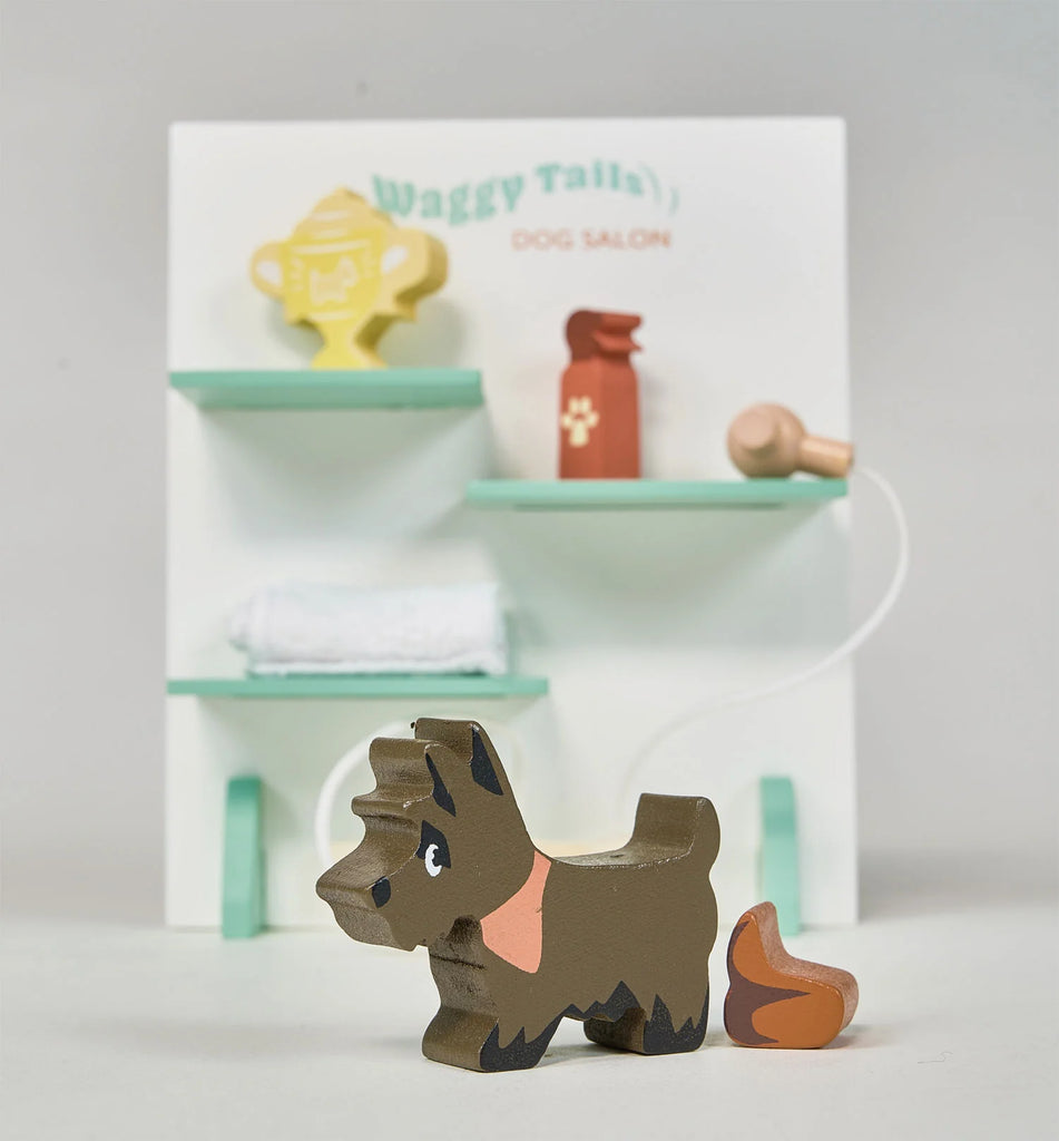 A small wooden dog figurine stands in front of a backdrop labeled "Waggy Tails Dog Salon." The backdrop, part of the Waggy Tails Dog Salon, includes two shelves holding a trophy, a red spray bottle, and a rolled-up towel. The brown figurine features seven breeds of dogs, each with unique detailing.