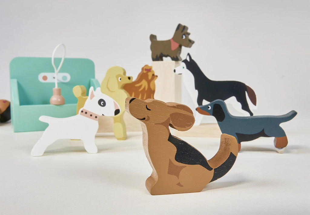 A set of colorful wooden toy dogs in 7 breeds and poses, part of the Waggy Tails Dog Salon play set, are displayed on a white background. A miniature light blue chair and some small toy accessories are visible in the background.