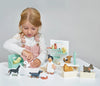 A young girl with blonde hair, wearing a white shirt and pink overalls, enjoys her Waggy Tails Dog Salon. The set features 7 breeds of dogs, salon furniture, and a groomer character. Everything is neatly arranged on a white table with a gray background.