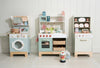 Three pastel-colored Kitchen Dresser and Dishwasher wooden toy kitchen sets—including a children's play kitchen with a washing machine, stove, oven, sink, toy dishwasher, shelves with play food, dishes, baskets, and accessories—all arranged against a white-paneled wall on a tiled floor—create the perfect pretend cooking space.