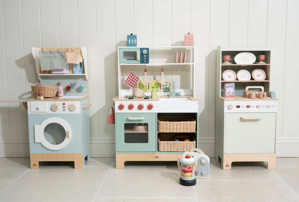 Three pastel-colored Kitchen Dresser and Dishwasher wooden toy kitchen sets—including a children's play kitchen with a washing machine, stove, oven, sink, toy dishwasher, shelves with play food, dishes, baskets, and accessories—all arranged against a white-paneled wall on a tiled floor—create the perfect pretend cooking space.