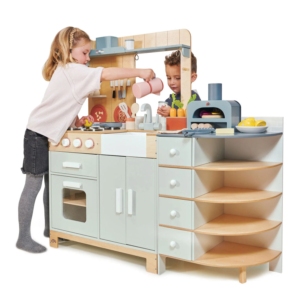 Two young children play together in the La Fiamma Grand Kitchen, pretending to cook with toy food and utensils. This kids kitchen set features shelves, a pizza oven toy, an oven, a sink, and several playful accessories.