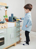 A young boy in a light blue shirt and dark pants plays with the La Fiamma Grand Kitchen, pretending to cook food on the stove of his kids kitchen set.