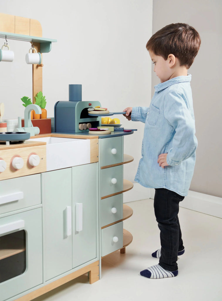 A young boy in a light blue shirt and dark pants plays with the La Fiamma Grand Kitchen, pretending to cook food on the stove of his kids kitchen set.