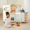 A young girl sits on the floor playing with toy fruits in front of the La Fiamma Grand Kitchen, a kids kitchen set featuring pastel colors, a wooden play kitchen design, and various kitchen accessories.
