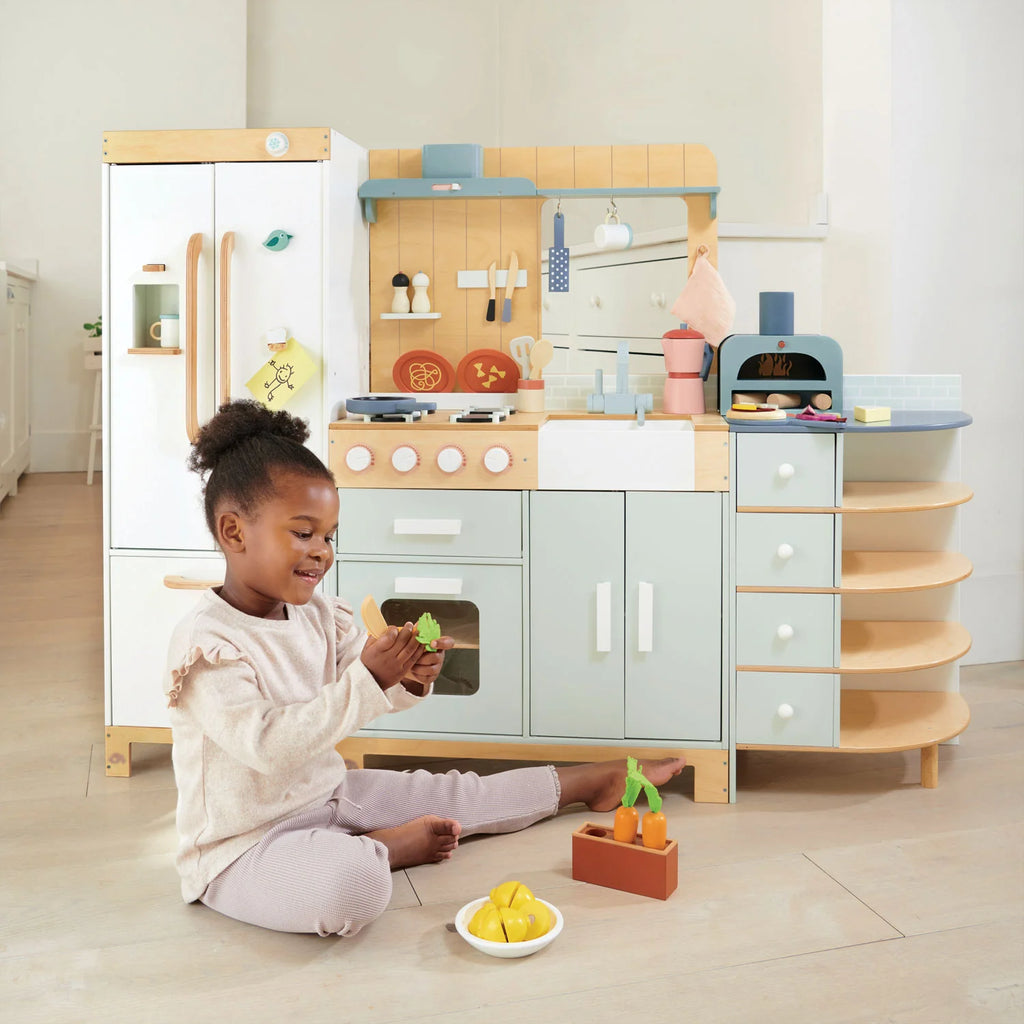 A young girl sits on the floor playing with toy fruits in front of the La Fiamma Grand Kitchen, a kids kitchen set featuring pastel colors, a wooden play kitchen design, and various kitchen accessories.
